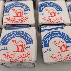 Puede incluir: Barras de jabón blancas con etiquetas azules y rojas que muestran una ilustración de camello y texto en árabe. El embalaje consistente resalta la marca del producto.