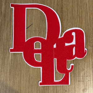May include: A red embroidered patch with the letters "DeLe" in a stylized font. The letters are outlined in white, and the patch is set against a wooden background. The patch is likely for clothing or accessories.