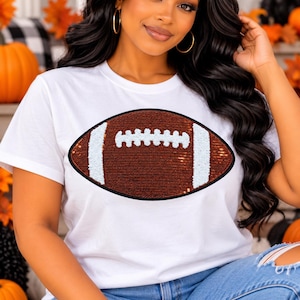 May include: A white short-sleeved t-shirt featuring a large, central graphic of an American football. The football design is embellished with brown and white sequins, creating a textured, shimmering appearance against the plain white fabric.