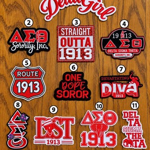 May include: Eleven embroidered patches with red, white, and black designs featuring Greek letters, the text Delta Girl, Straight Outta 1913, and elephant motifs, arranged above a purple ruler measuring 13 inches in length on a wood grain surface.