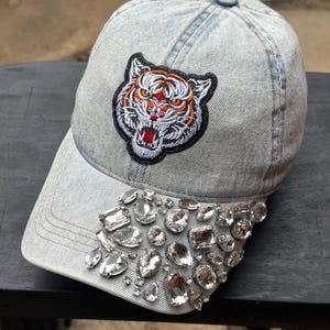 May include: Light blue denim baseball cap with a tiger patch and sparkling crystal embellishments along the brim. The tiger patch features orange, white, and black details. A stylish accessory.