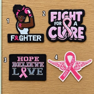 May include: Four embroidered patches featuring breast cancer awareness themes. Patch 1 shows a person with a pink boxing glove and the text F&GHTER. Patch 2 says FIGHT FOR A CURE with a pink ribbon. Patch 3 says HOPE BELIEVE LOVE. Patch 4 is a winged pink ribbon.