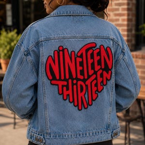 May include: A blue denim jacket featuring a large, red, heart-shaped patch on the back. The patch contains the black-outlined text "NINETEEN THIRTEEN" in a bold, stylized font. The jacket has a classic collar, long sleeves, and buttoned cuffs.