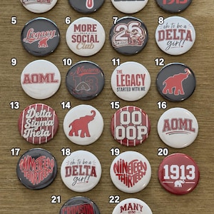 May include: A collection of twenty-two circular pinback buttons featuring various designs in red, white, and black. The buttons display text such as 'Delta Sigma Theta', '1913', 'Legacy', 'Diva', and 'AOML', along with elephant graphics and striped patterns.