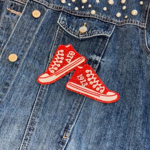 May include: A denim jacket with pearl embellishments and two red sneaker patches. The patches have white accents and the text "ΔΣΘ 1913". The jacket has button closures and pockets.