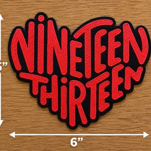 May include: A heart-shaped embroidered patch featuring the red text NINETEEN THIRTEEN on a black background. The patch measures 5 inches in height and 6 inches in width as indicated by the white arrows and measurement text on the image.
