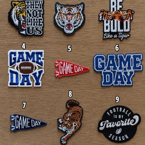 May include: A collection of nine embroidered patches featuring tiger motifs, football imagery, and text including 'THEY NOT LIKE US', 'BE BOLD Like a Tiger', 'GAME DAY', and 'FOOTBALL IS MY FAVORITE SEASON' in various colors like blue, white, red, orange, and black.