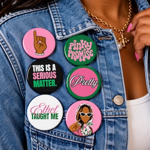 May include: Six circular pinback buttons attached to a blue denim jacket. Designs include a pink button with a hand gesture, a green button with the text Pinky Promise, a black button with the text This Is A Serious Matter, a pink and green button with the word Pretty, a white button with the text Ethel Taught Me, and a pink button featuring an illustration of a person wearing a headscarf and sunglasses.