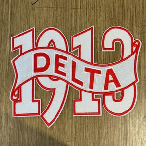 May include: A white and red embroidered patch featuring the number "1913" with the word "DELTA" on a banner. The patch has a white background with red outlining and lettering. The design is likely for a collegiate or Greek organization.