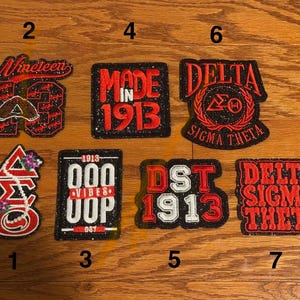 May include: A collection of seven embroidered patches with black glitter borders. The patches feature red and white text and symbols, including Greek letters, numbers, and phrases like "Made in 1913" and "Delta Sigma Theta."