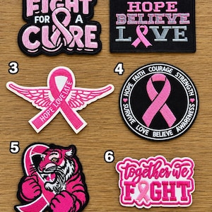 May include: Six embroidered patches featuring pink awareness ribbons. Patch 1 says FIGHT FOR A CURE. Patch 2 says HOPE BELIEVE LOVE. Patch 3 is a ribbon with wings. Patch 4 is circular with text. Patch 5 features a tiger. Patch 6 says together we FIGHT.