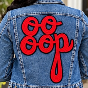 May include: A blue denim jacket featuring a large, red, embroidered patch on the back. The patch displays the text "OO OOP" in a stylized, cursive font with a thick black border. The jacket has visible stitching, a collar, and metal buttons.