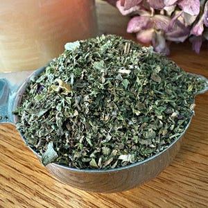 May include: A close-up of a metal measuring cup filled with dried herbs. The herbs are a mix of green and brown, and they appear to be finely chopped.