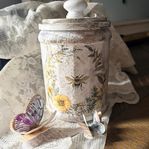 May include: A cylindrical jar with a distressed white finish, featuring a lid with a white knob. The jar is decorated with botanical illustrations of yellow flowers, green foliage, and a bee. Two paper butterfly cutouts sit in front of the jar.
