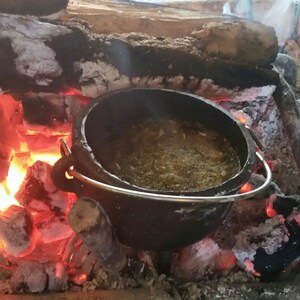 May include: A black cast iron pot with a metal handle sits on top of hot coals in a fire pit. The pot is filled with a brown liquid and has a layer of white foam on top.
