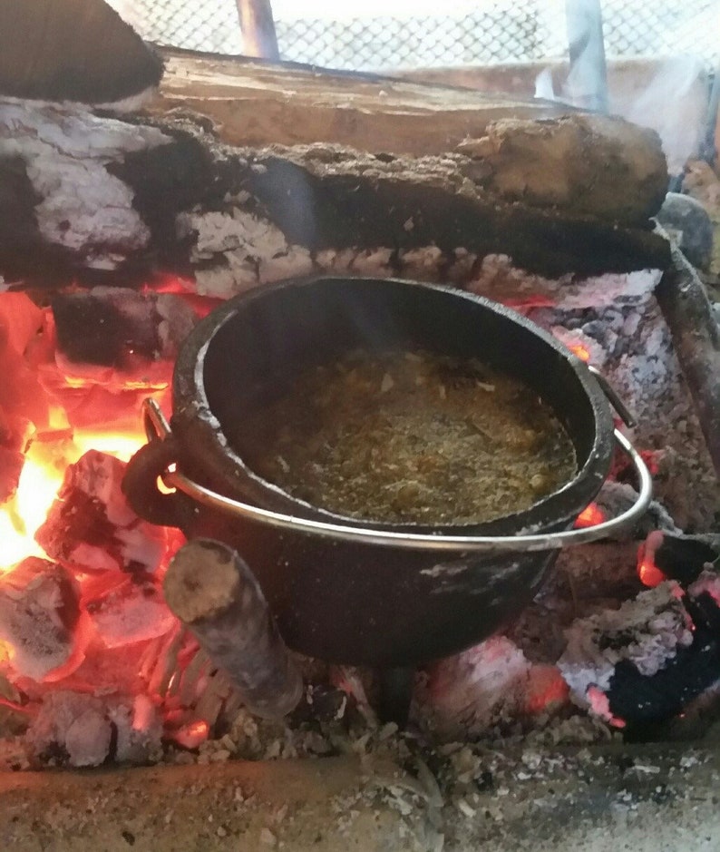 May include: A black cast iron pot with a metal handle sits on top of hot coals in a fire pit. The pot is filled with a brown liquid and has a layer of white foam on top.