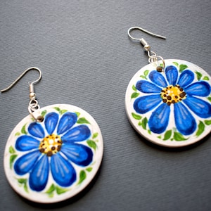 Puede incluir: Un par de pendientes con diseños florales azules y verdes sobre bases redondas de cerámica blanca. Los pendientes tienen ganchos plateados.