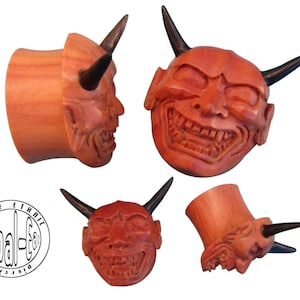 May include: Set of four wooden ear plugs, each carved with a grinning devil face and dark horns. The plugs are reddish-brown and feature detailed facial expressions. The brand logo "tribal-ea" is visible in the lower left corner.