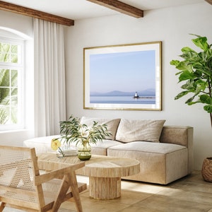 May include: A light and airy living room with a framed landscape print of a lighthouse. The room includes a beige sofa, a wooden coffee table, and a rattan chair. A large potted plant and white curtains are also visible.