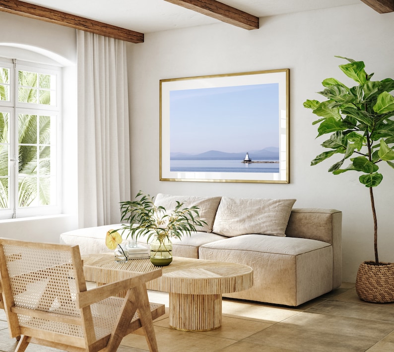 May include: A light and airy living room with a framed landscape print of a lighthouse. The room includes a beige sofa, a wooden coffee table, and a rattan chair. A large potted plant and white curtains are also visible.