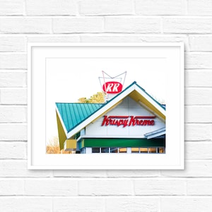 May include: A white building with a green roof and a red and white sign that says "Krispy Kreme" in red letters. The sign is on the front of the building and there is a red and white sign with the letters "KK" on top of the building.