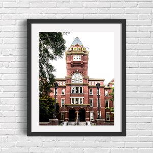 May include: A framed photograph of a red brick building with a tall tower and a clock face. The building has a white trim and a large arched entrance. The building is surrounded by trees.