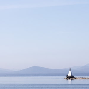 May include: A tranquil coastal scene with a white lighthouse and black railing on a stone pier. The calm, blue water mirrors the sky and distant, misty mountains. The image conveys a sense of peace and serenity.