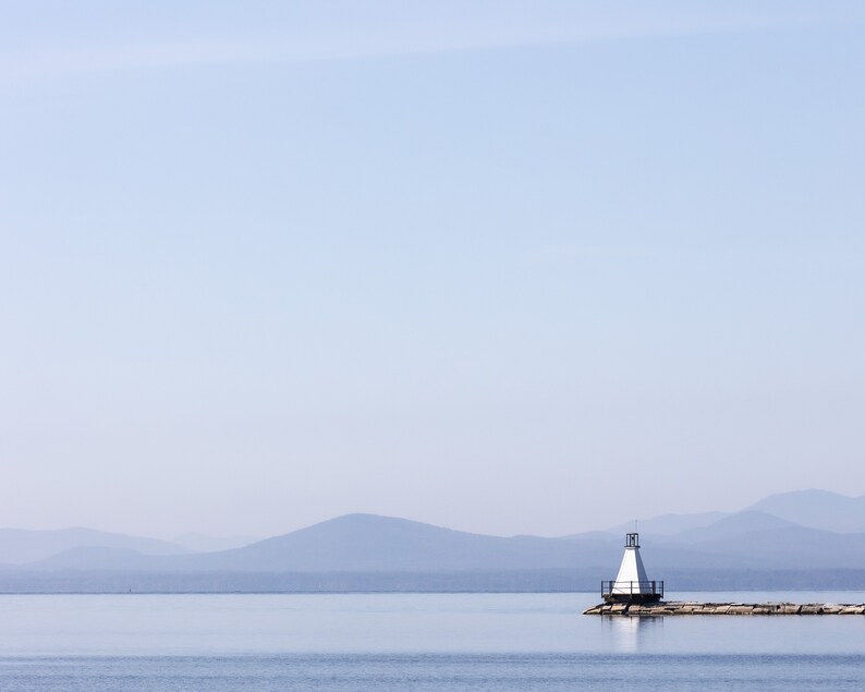 May include: A tranquil coastal scene with a white lighthouse and black railing on a stone pier. The calm, blue water mirrors the sky and distant, misty mountains. The image conveys a sense of peace and serenity.