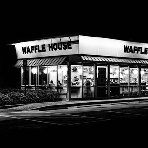 May include: A black and white photograph of a Waffle House restaurant at night. The sign above the entrance reads "Waffle House" and "Waffle". The restaurant has large windows and is lit up inside.
