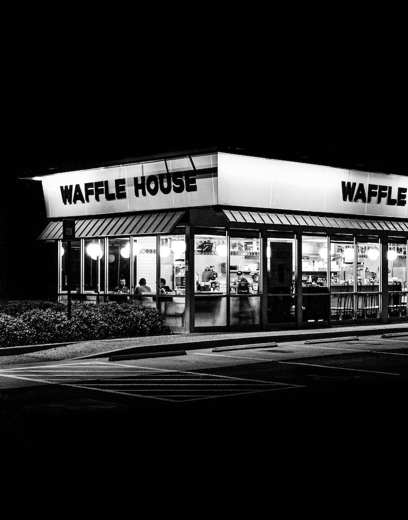 May include: A black and white photograph of a Waffle House restaurant at night. The sign above the entrance reads "Waffle House" and "Waffle". The restaurant has large windows and is lit up inside.