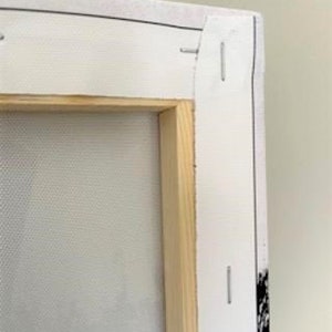 May include: Close-up of the back of a stretched canvas painting. The canvas is wrapped around a wooden frame and secured with staples. The canvas is white and the frame is natural wood.