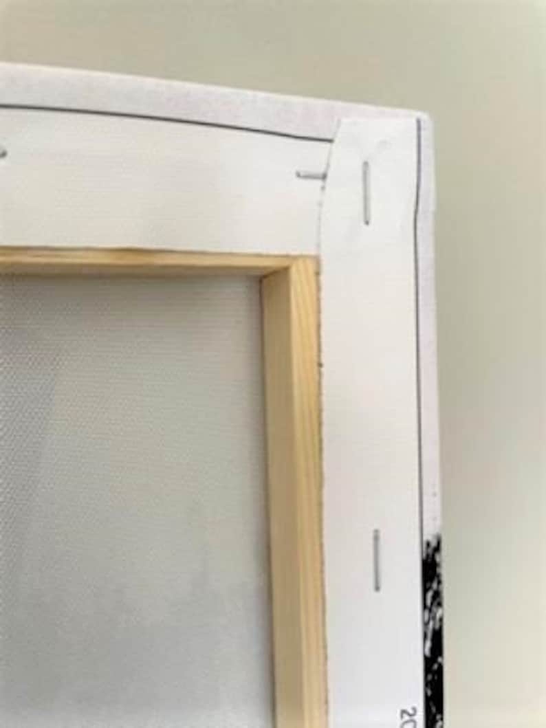 May include: Close-up of the back of a stretched canvas painting. The canvas is wrapped around a wooden frame and secured with staples. The canvas is white and the frame is natural wood.