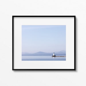 May include: Framed print of a coastal scene with a white lighthouse with a black top on a stone pier. The image shows calm, blue water, distant mountains, and a pale blue sky, creating a tranquil landscape. The frame is black.