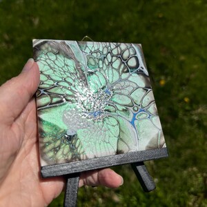 May include: A small, square abstract art piece with a fluid design in shades of green, silver, and blue, mounted on a miniature black easel. The artwork features a unique, organic pattern, perfect for home decor.