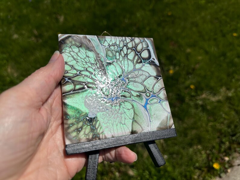 May include: A small, square abstract art piece with a fluid design in shades of green, silver, and blue, mounted on a miniature black easel. The artwork features a unique, organic pattern, perfect for home decor.