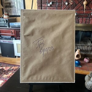 May include: A framed artwork with a brown paper backing, displayed on a black easel. The paper has a signature and the date 1/14/23 in black ink. The artwork is set against a domestic interior background.