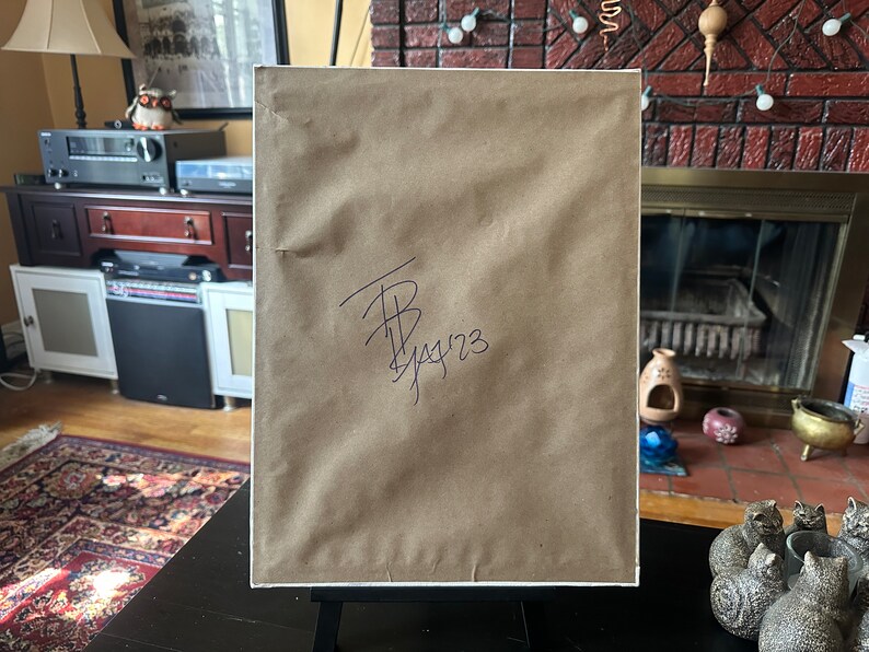 May include: A framed artwork with a brown paper backing, displayed on a black easel. The paper has a signature and the date 1/14/23 in black ink. The artwork is set against a domestic interior background.