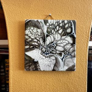 May include: Square abstract art piece with a black, white, and gray pattern resembling a flower or cell structure. The artwork is mounted on a yellow wall with a gold-colored hook.