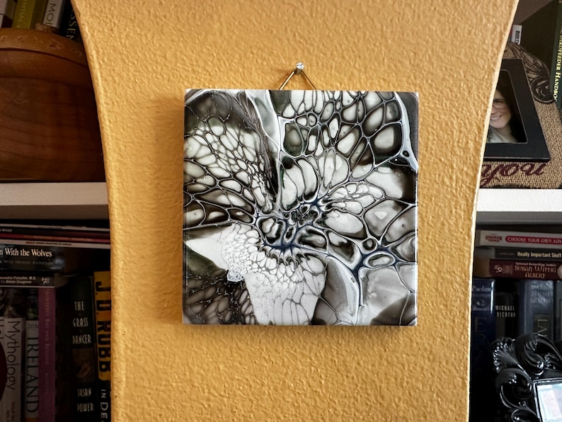 May include: Square abstract art piece with a black, white, and gray pattern resembling a flower or cell structure. The artwork is mounted on a yellow wall with a gold-colored hook.