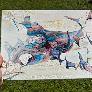 May include: Abstract acrylic pour painting on canvas. The artwork features a cream-colored background with flowing patterns in shades of blue, red, gold, and gray. The fluid art style creates a unique and textured visual effect.