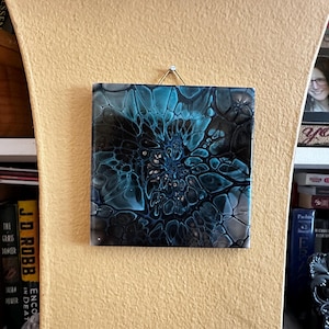 May include: A square wall art piece featuring an abstract acrylic pour design with swirling patterns of deep black, teal, and white. The surface has a glossy finish and is suspended by a thin metallic wire attached to the top edge of the tile.