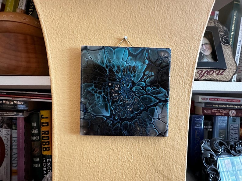 May include: A square wall art piece featuring an abstract acrylic pour design with swirling patterns of deep black, teal, and white. The surface has a glossy finish and is suspended by a thin metallic wire attached to the top edge of the tile.