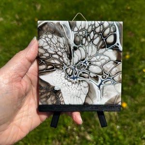 May include: Abstract art piece on a small black easel. The artwork features a fluid design with black, white, and gray tones. The painting has a unique, organic pattern, and is displayed on a miniature wooden easel.