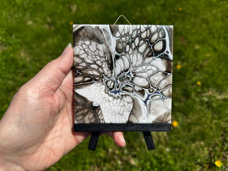 May include: Abstract art piece on a small black easel. The artwork features a fluid design with black, white, and gray tones. The painting has a unique, organic pattern, and is displayed on a miniature wooden easel.