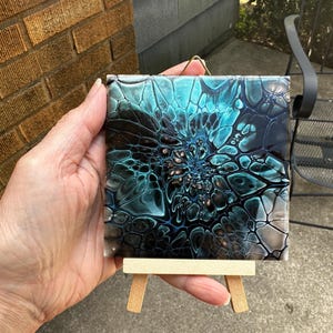 May include: A square tile featuring an abstract fluid acrylic pour design with swirling patterns of deep black, vibrant teal, and metallic copper. The tile is displayed on a small, light-colored wooden easel and includes a gold wire hanger.