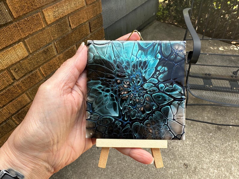 May include: A square tile featuring an abstract fluid acrylic pour design with swirling patterns of deep black, vibrant teal, and metallic copper. The tile is displayed on a small, light-colored wooden easel and includes a gold wire hanger.