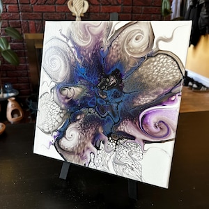 Puede incluir: Un cuadro cuadrado sobre lienzo con un dise&ntilde;o abstracto de vertido de acr&iacute;lico. La composici&oacute;n muestra un n&uacute;cleo central en azul oscuro y negro que se irradia hacia patrones arremolinados de blanco, plata met&aacute;lica y morado intenso con celdas.