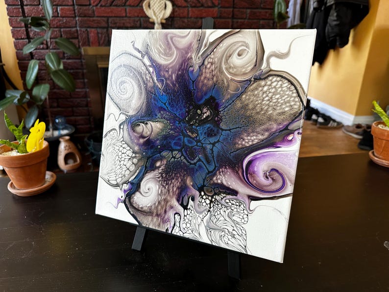 Puede incluir: Un cuadro cuadrado sobre lienzo con un dise&ntilde;o abstracto de vertido de acr&iacute;lico. La composici&oacute;n muestra un n&uacute;cleo central en azul oscuro y negro que se irradia hacia patrones arremolinados de blanco, plata met&aacute;lica y morado intenso con celdas.
