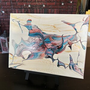 May include: Abstract acrylic painting on canvas. The artwork features a cream-colored background with flowing shapes in shades of blue, pink, and black. Gold accents add detail. The canvas is displayed on a small black easel.