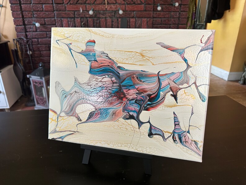 May include: Abstract acrylic painting on canvas. The artwork features a cream-colored background with flowing shapes in shades of blue, pink, and black. Gold accents add detail. The canvas is displayed on a small black easel.
